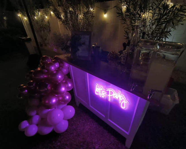 A dimly lit outdoor party scene features a bar table adorned with a glowing "Let's Party" sign and a cluster of pink balloons beside it.