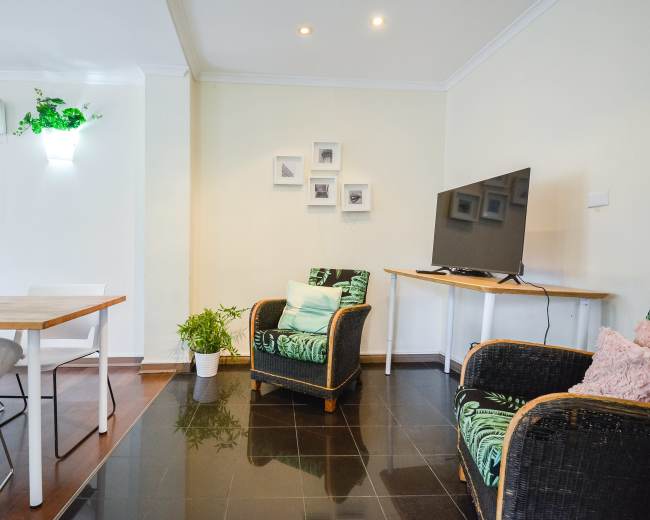 The image shows a cozy living area with a wooden table, two chairs, and a television mounted on the wall, complemented by potted plants and framed pictures.