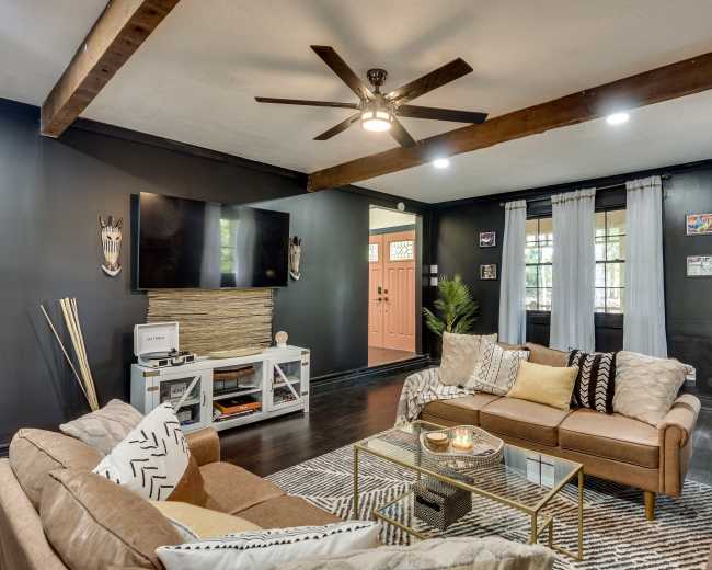 A modern living room features two sofas, a coffee table, a mounted television, and a decorative wall with wood beams and a door leading outside.