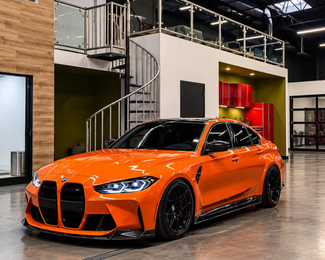 A bright orange BMW M3 is parked inside a modern industrial-style interior featuring wood paneling and a spiral staircase.