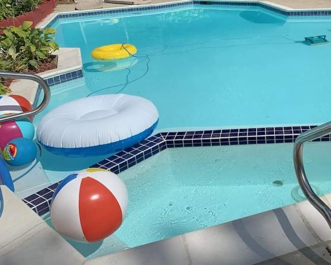 A swimming pool features colorful beach balls and inflatable floats, with a robotic cleaner in the water and steps leading into the pool.