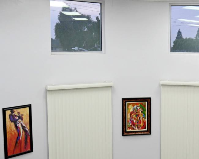 A corner of a room with two large windows, white blinds, and four framed paintings on the walls.