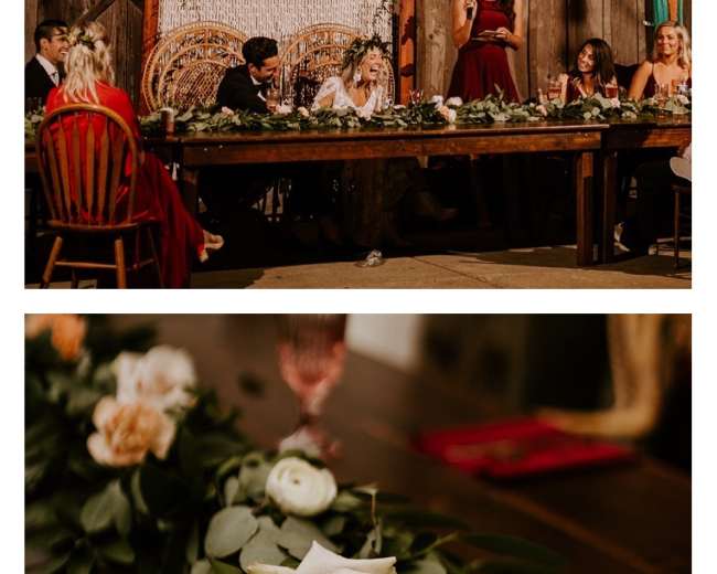 A woman stands and speaks at a long banquet table adorned with greenery and flowers in a rustic, wooden venue.
