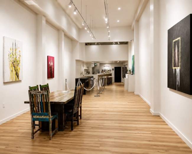 The image depicts a spacious art gallery with wooden floors, featuring paintings on the walls and a large wooden table with colorful chairs.