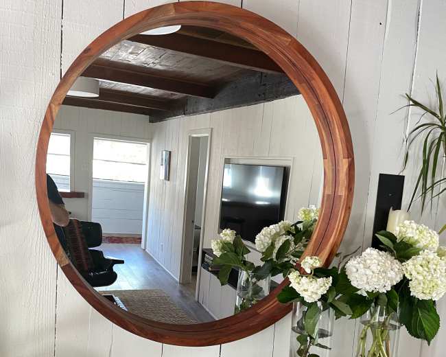 A round wooden mirror reflects a room with a piano, greenery, and light-colored walls.