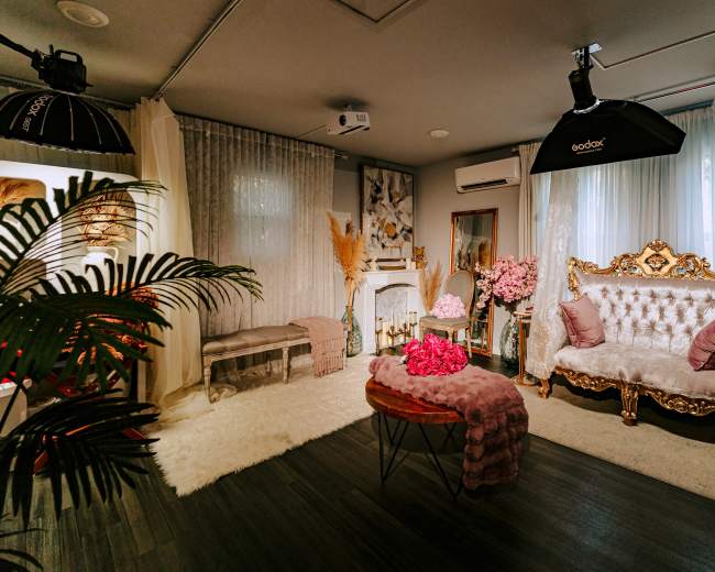 The image shows a stylish interior living space featuring a gold-accented sofa, a plush rug, and decorative plants, with soft lighting and draped curtains.