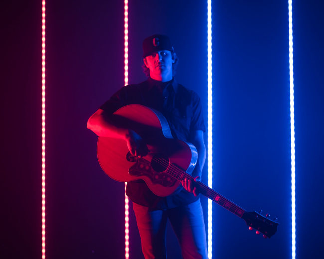 A musician stands holding an acoustic guitar in front of vertical light strips in red and blue.