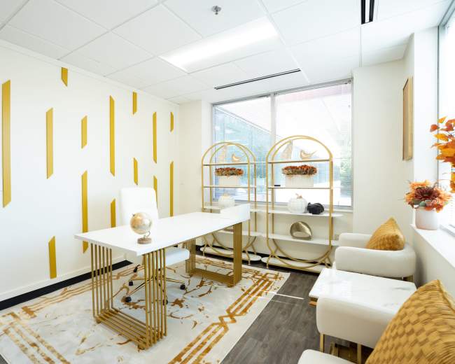 The image shows a modern office space featuring a white table, two white chairs, a decorative bookshelf, and a rug with a geometric pattern against a backdrop of a gold-striped wall.