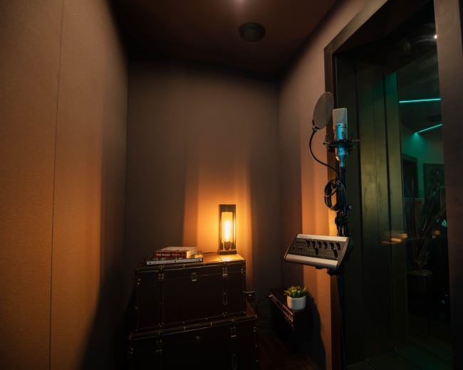 The image shows a small recording booth with a microphone, music equipment, a lamp, and a plant in a corner.