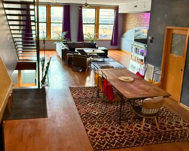 The image shows a spacious living area with a dining table, a staircase, and large windows that allow natural light to enter.