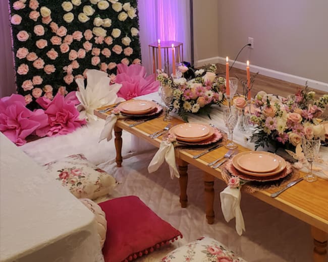 A decorated dining area with a low wooden table set for a meal, surrounded by floral arrangements and cushions on the floor.