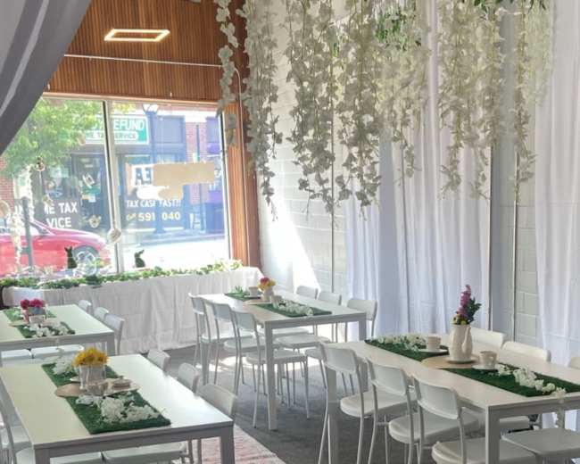 The image shows a bright indoor space with several white tables set with green table runners and floral decorations, under a canopy of hanging greenery.
