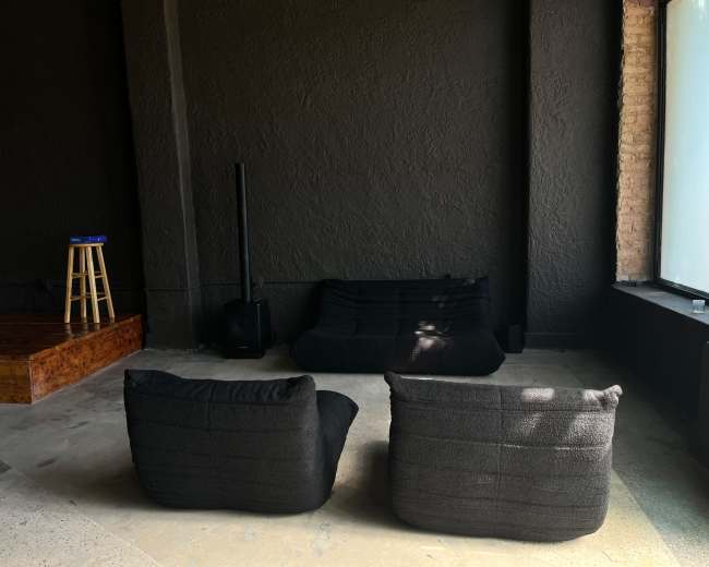 The image shows a minimalist room featuring two black lounge chairs facing a low black sofa, with a wooden stage in the background and a large window.