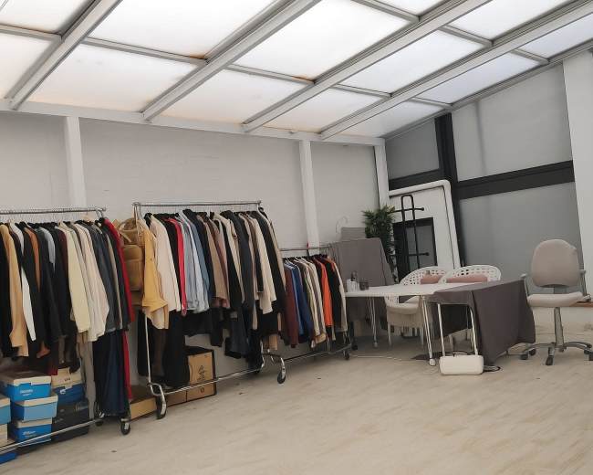 The image shows a spacious room with a rack of clothing on one side and a table with chairs on the other.