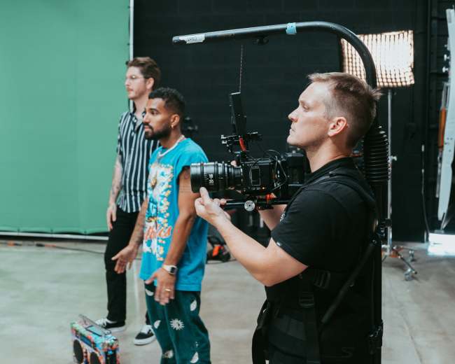 A camera operator stands in front of two individuals in a studio with a green screen backdrop.