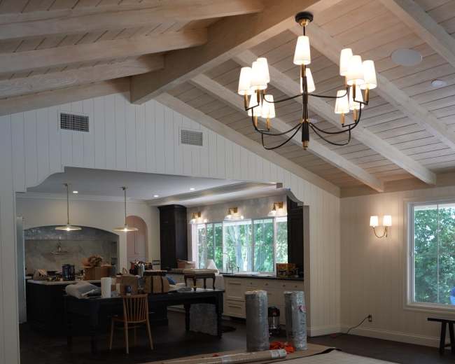 The image shows a spacious living area with a dining table, modern kitchen, and large windows, featuring exposed wooden beams and a patterned rug on the floor.