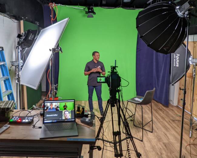 A person stands in front of a green backdrop in a studio, with lighting equipment and a laptop displaying a video call setup on a table.