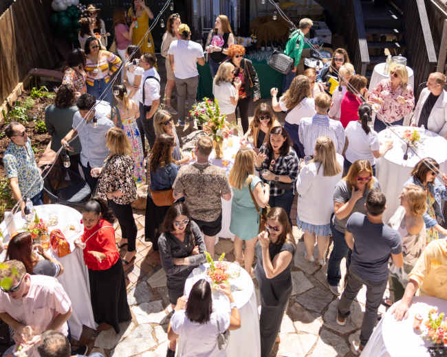 A large outdoor gathering takes place in a backyard, featuring numerous guests mingling around tables adorned with floral arrangements.