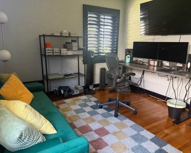 The image shows a neatly arranged home office with a green sofa, a desk featuring two monitors, and a shelf with various electronic devices and a floor lamp.