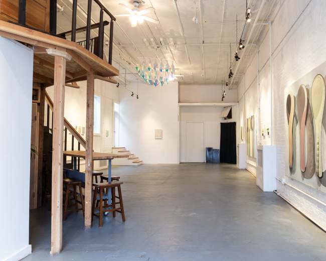 The image depicts a spacious art gallery with high ceilings, featuring a staircase, modern artworks on the walls, and a minimalist, uncluttered floor layout.
