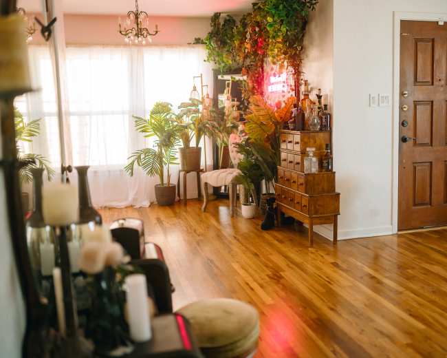 A well-lit room featuring wooden flooring, a large window with sheer curtains, various indoor plants, a wooden cabinet, and a decorative wall with neon lights.