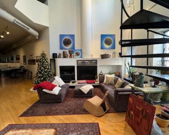 The image shows a spacious living room featuring a large sectional sofa, a coffee table, and a spiral staircase, with a Christmas tree in the corner and various artworks hanging on the walls.
