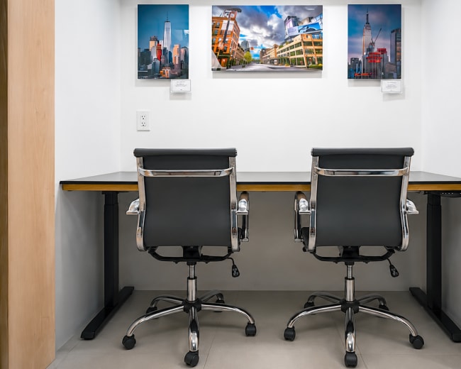 The image shows a small workspace featuring a modern desk with two black office chairs and three framed photographs on the wall above.