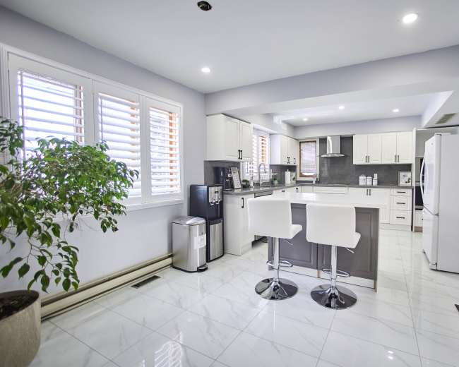 The image shows a modern kitchen featuring white cabinets, a gray island with bar stools, and large windows with shutter blinds.