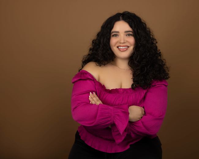 A woman with curly hair is sitting and smiling, wearing a pink off-shoulder top against a brown background.