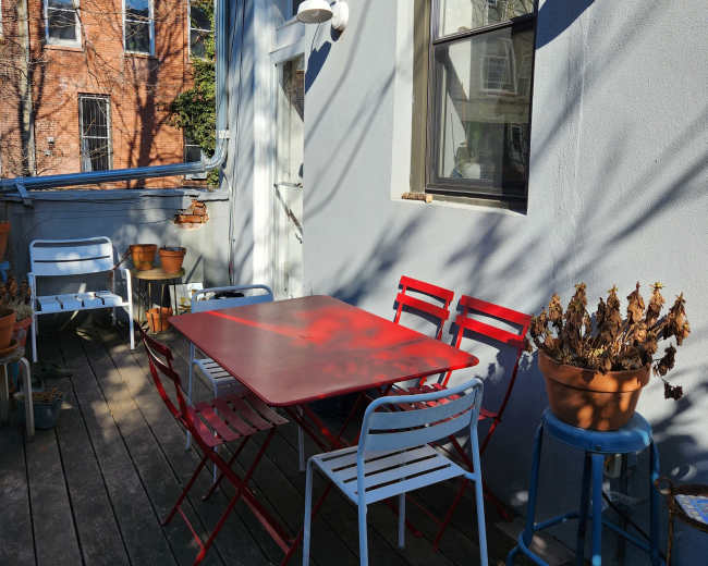 A red table is surrounded by red and white chairs on a wooden deck, with potted plants and a light fixture attached to the gray wall.