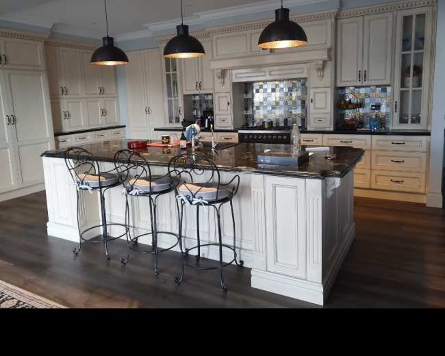 The image shows a kitchen with a central island featuring bar stools and pendant lights, wooden flooring, and cabinetry in a light color.