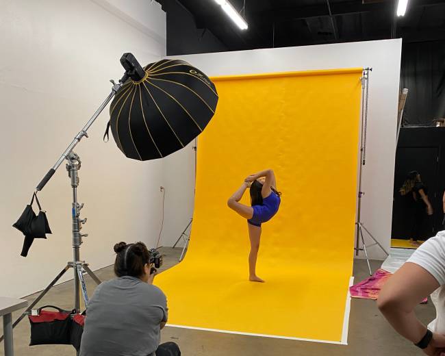 A dancer performs a pose on one leg in front of a yellow backdrop as a photographer captures the image.