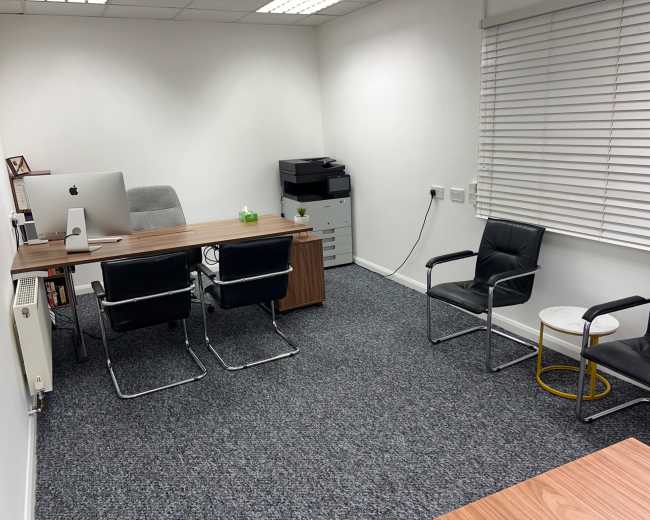The image shows a minimalist office space with a wooden desk, an Apple computer, two black chairs, a small side table, and a printer against a white wall with blinds.