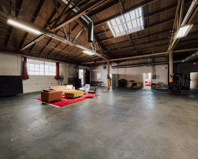 The image shows a spacious, industrial-style room with high wooden ceilings, large windows, and a seating area centered on a red rug.