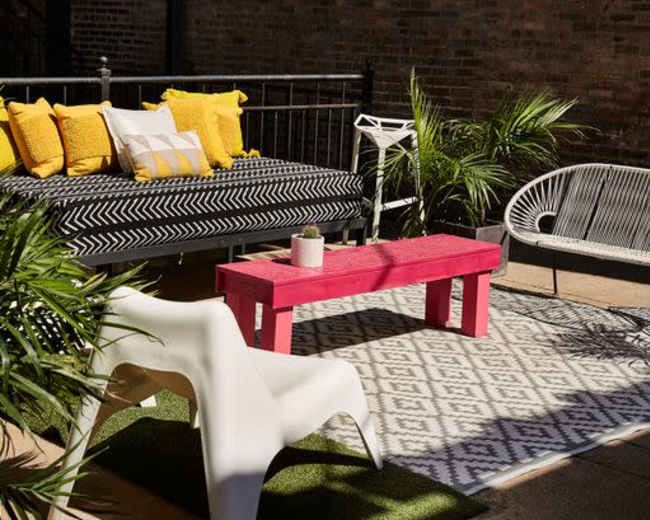 The image shows a contemporary outdoor seating area featuring a black and white striped sofa with yellow pillows, a pink coffee table, and a patterned rug surrounded by potted plants.