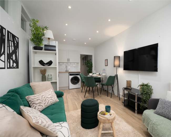 The image shows a modern living room featuring green sofas, a dining area with four chairs, and a wall-mounted television.