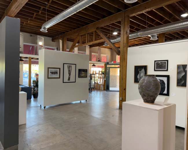 The image shows an art gallery interior with framed artwork displayed on the walls and a ceramic vase on a pedestal in the foreground.