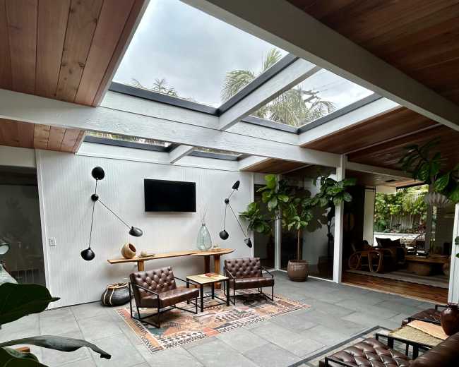 The image depicts a modern living space with a skylight, featuring two leather chairs, a coffee table, and various indoor plants.