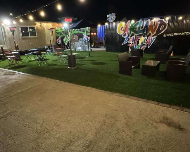An outdoor seating area with string lights and decorative murals, featuring grass-like flooring and a mix of tables and chairs.