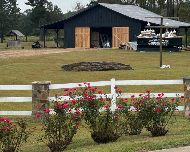 A dark barn with wooden doors stands in a grassy area surrounded by trees and a white fence, with ornamental decorations hanging nearby.