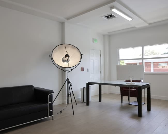 The image shows a minimalist office space with a black leather couch, a desk with a chair, and a large floor lamp.