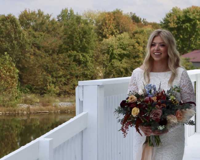 A woman in a white lace wedding dress walks along a white railing by a body of water, holding a bouquet of flowers.