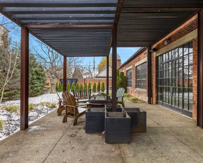The image depicts a patio area with a covered seating arrangement, including wooden chairs and metal planters, surrounded by a landscaped yard.