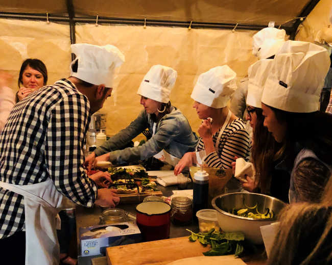 A group of people wearing chef hats prepares food in a tented kitchen.