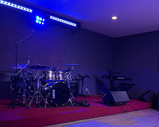 A drum set is positioned on a stage with red carpeting, accompanied by a keyboard and blue lighting illuminating the scene.