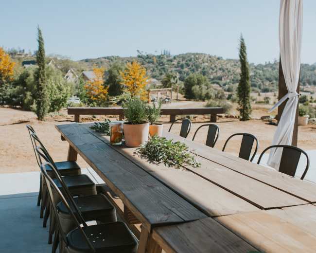 A long wooden table with black chairs is set outside, surrounded by greenery and a rustic landscape in the background.