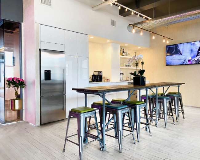 A modern kitchen features a long wooden table with high stools, a stainless steel refrigerator, and a wall-mounted television.
