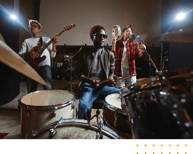 Three musicians perform together in a recording studio, with one playing the drums, another on bass, and a third singing into a microphone.