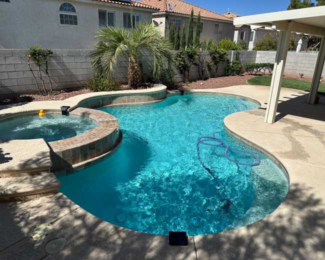 The image shows a swimming pool and hot tub in a backyard surrounded by a concrete patio and landscaped area.