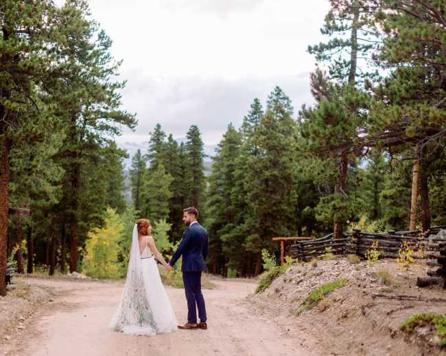 A couple stands hand in hand on a dirt path surrounded by tall pine trees.